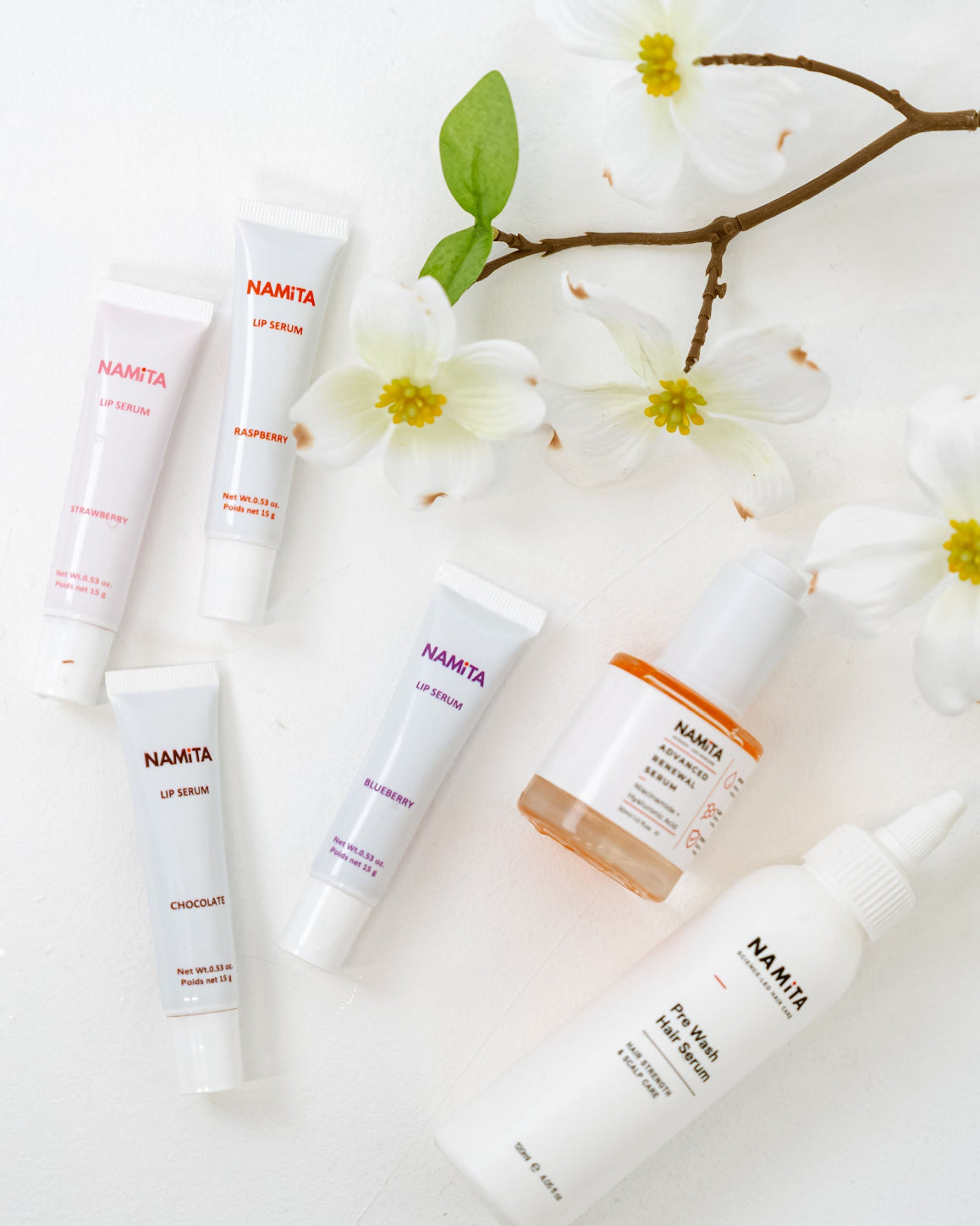A flat-lay arrangement of six NAMTA skincare products on a white background, styled with white magnolia flowers and a green leaf. The products include lip balm, moisturizer, face wash, and serum in white tubes and bottles with pink, purple, and orange accents.
